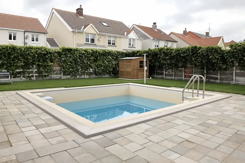 Piscine coque enterrée avec échelle inox et plage dallée claire dans un jardin de lotissement, haie et abri de jardin au fond