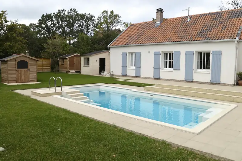 Piscine coque avec escalier d’angle, plage claire et abri de jardin en bois au fond de la pelouse