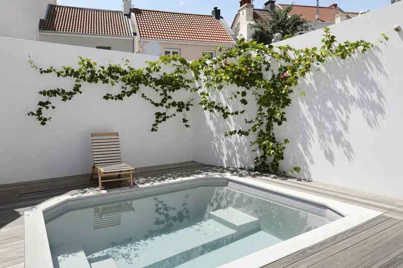 Mini piscine aux angles coupés encastrée dans une terrasse en bois, au pied de murs blancs couverts d’une plante grimpante, dans une cour de ville