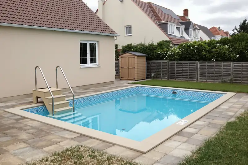 Piscine coque de 6 mètres sur 3 avec escalier et échelle inox, plage dallée, devant un pavillon beige de lotissement