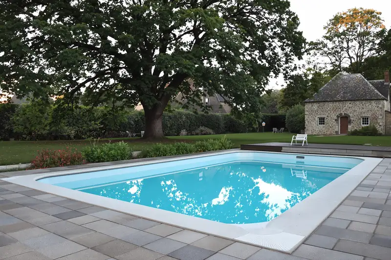 Piscine coque de 8 mètres sur 4 à margelles claires et plage dallée grise, grand chêne et dépendance en pierre au fond