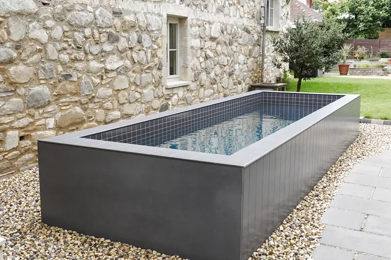 Mini bassin maçonné surélevé revêtu de carreaux gris foncé dans la cour d’une maison en pierre