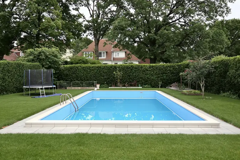 Piscine rectangulaire avec échelle inox au milieu d’une pelouse bordée de haies et de grands arbres, maison à toit de tuiles en arrière-plan