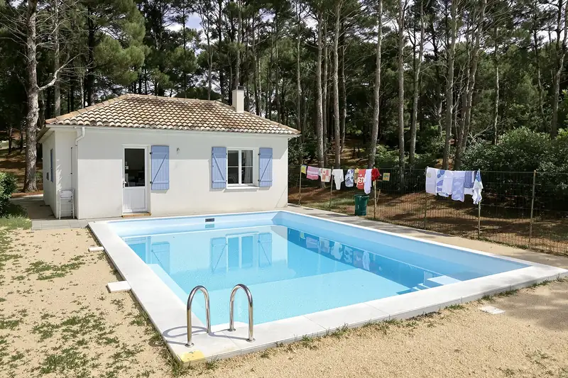 Piscine rectangulaire avec échelle inox devant une maison basse aux volets bleus, sous de grands pins, sur un sol de sable et d’aiguilles
