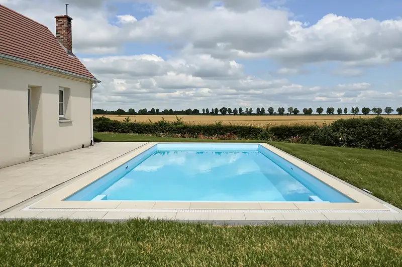 Piscine rectangulaire à margelles claires au bord d’une pelouse, face à un champ de céréales et à une rangée d’arbres, à côté d’une maison crépie au toit de tuiles