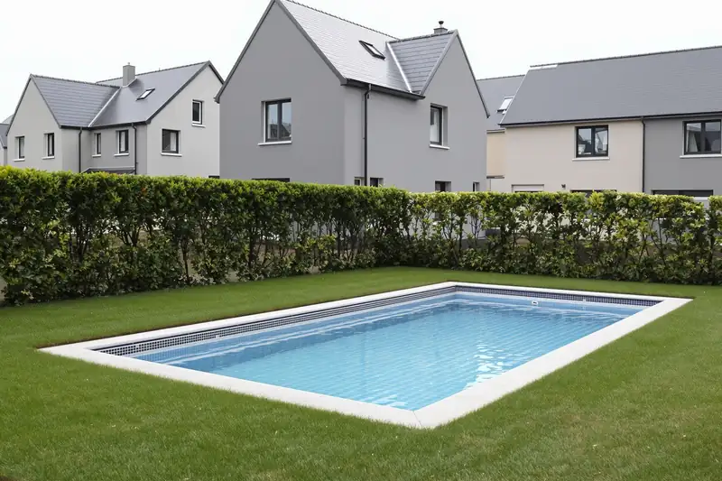 Piscine rectangulaire enterrée dans la pelouse d’une maison neuve, derrière une haie, face à d’autres maisons récentes aux façades grises