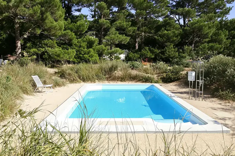 Piscine rectangulaire dans un jardin sablonneux planté de graminées, sous les pins maritimes, avec un transat blanc et une échelle de bain