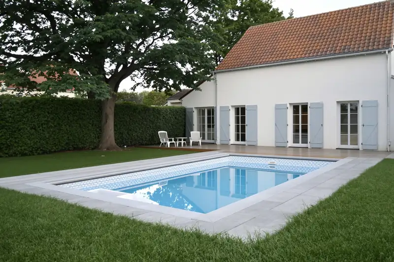 La même piscine rénovée, eau claire sur un revêtement bleu avec frise, margelles et plage en pierre claire