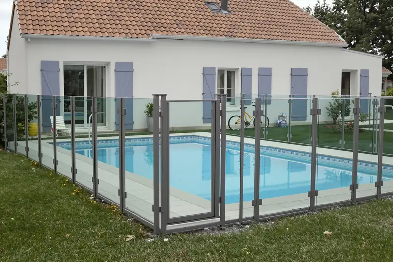 Barrière en panneaux de verre et poteaux gris autour d’une piscine enterrée, devant une maison aux volets bleus