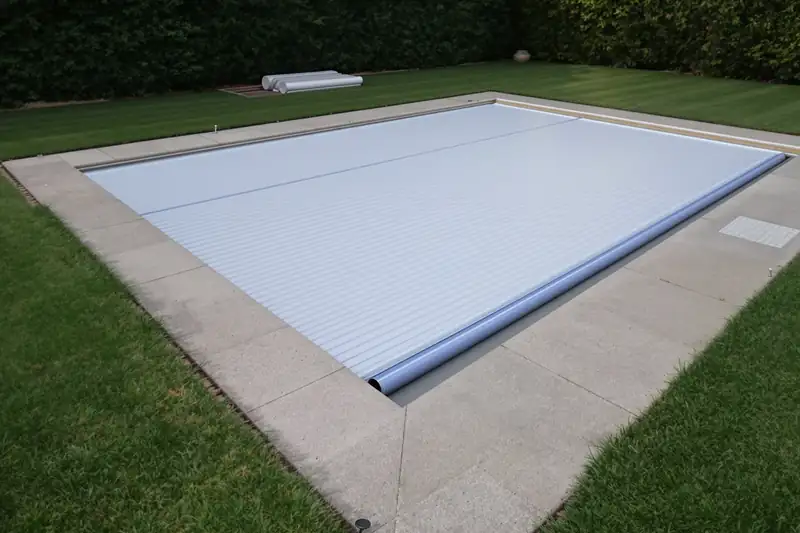 Volet de piscine à lames claires déroulé sur un bassin rectangulaire bordé de dalles et de pelouse