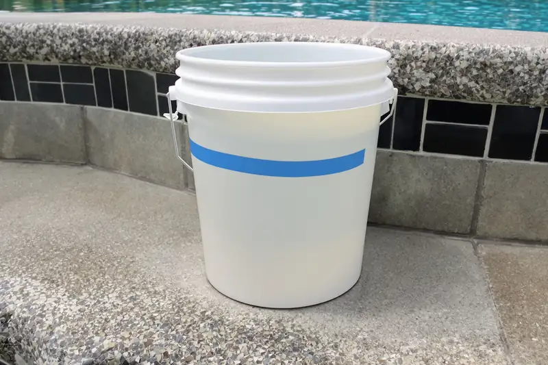 Seau blanc marqué d’un ruban bleu, posé au bord d’une piscine pour comparer la baisse du niveau d’eau