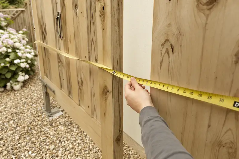 Des mains tendent un mètre ruban jaune en travers d’un portail en bois, au-dessus d’une allée gravillonnée